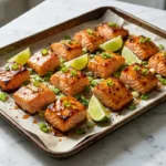 Hot honey lime salmon bites with caramelized edges, green onions, lime wedges, and honey glaze on a baking sheet