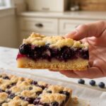 Hand holding a thick lemon blueberry crumble bar with shortbread base, jammy blueberry filling and golden crumble topping on white marble